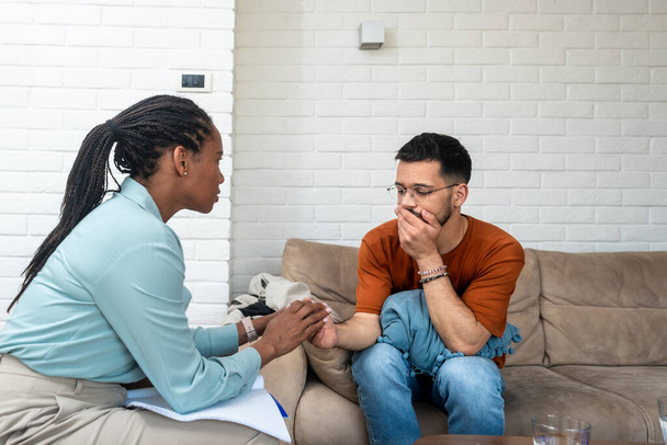 Young depressed man crying talking to mental health counselor during a session in the office. Stressed male at psychotherapy after divorce having emotional damage issue. Relationship difficulties. - Zdjęcie, obraz