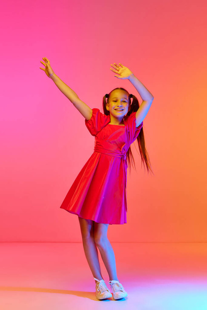 Charmant klein meisje in feestelijke jurk met paardenstaarten poseren over roze neon achtergrond. Concept van kindertijd, emoties, kindermode, dans, muziek. - Foto, afbeelding