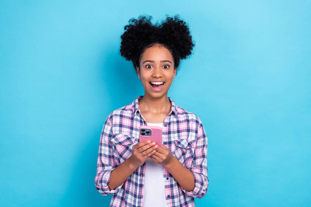 Photo of ecstatic girl perming coiffure dressed checkered shirt hold smartphone read impressive post isolated on blue color background. - Fotografie, Obrázek