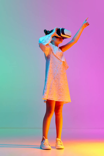Porträt eines kleinen Mädchens, das mit einer VR-Brille vor blaurosa Studiohintergrund in Neon steht. Bildung, Spaß, Spiel. Konzept der Kindheit, Emotionen, Lebensstil, Mode, Freude. Anzeige - Foto, Bild