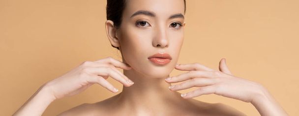 Asian woman with naked shoulders and natural makeup looking at camera isolated on beige, banner - Foto, imagen