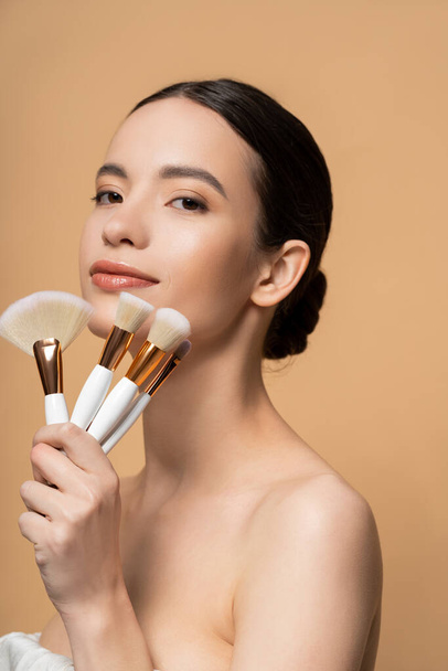 Asian woman with naked shoulders holding makeup brushes and looking at camera isolated on beige - Foto, afbeelding