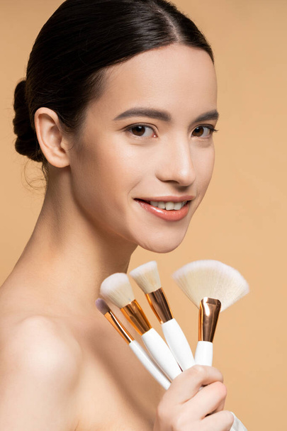 Portrait of smiling asian model holding makeup brushes and looking at camera isolated on beige - 写真・画像