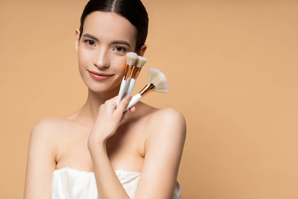 Pretty asian woman with naked shoulders in top holding makeup brushes and smiling isolated on beige - Фото, зображення