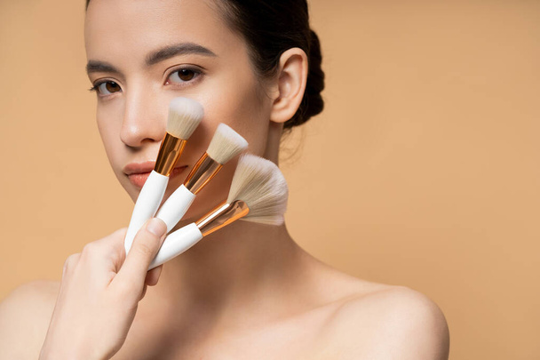 Young asian woman with naked shoulders holding makeup brushes near face isolated on beige - Фото, зображення