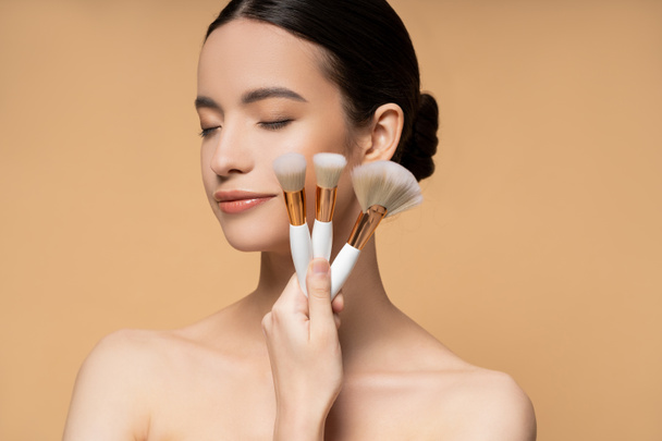 Young asian woman with closed eyes and naked shoulders holding makeup brushes isolated on beige - Zdjęcie, obraz