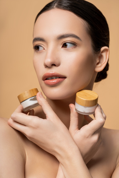 Pretty young asian woman with naked shoulders holding jars with cosmetic cream isolated on beige - Photo, Image