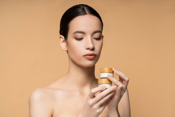 Brunette asian woman with naked shoulders holding jars with cosmetic cream isolated on beige - Foto, imagen
