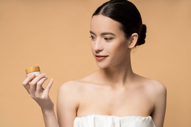 Positive young asian woman in top holding cosmetic cream while standing isolated on beige - Φωτογραφία, εικόνα