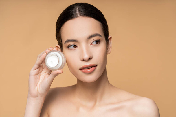 Young brunette woman with perfect skin and naked shoulders holding cosmetic cream isolated on beige - 写真・画像
