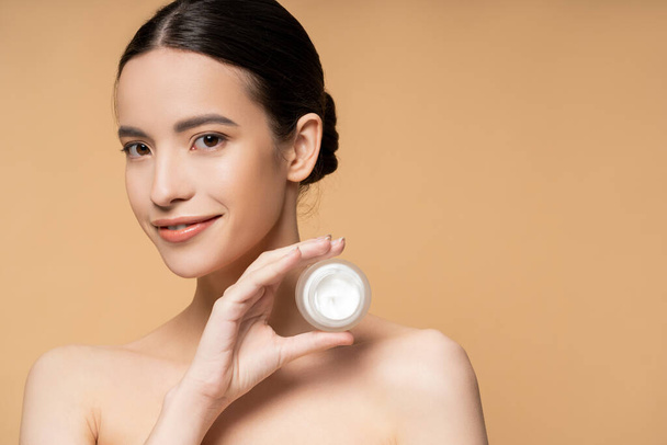 Happy asian woman with naked shoulders and natural makeup holding cosmetic cream isolated on beige - Zdjęcie, obraz