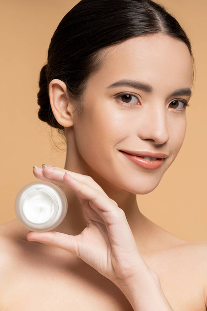 Positieve aziatische vrouw met perfecte huid houden moisturizer en kijken naar camera geïsoleerd op beige - Foto, afbeelding