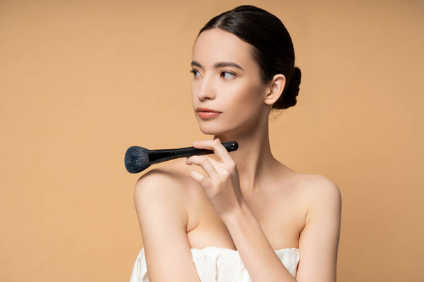 Young asian woman in top holding blush brush near naked shoulders isolated on beige - Фото, зображення