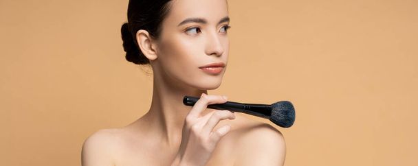Asian woman with perfect skin holding cosmetic brush near naked shoulder isolated on beige, banner - Foto, Imagen
