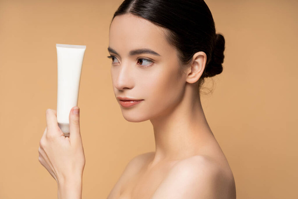 Young asian model with naked shoulders holding cosmetic lotion while posing isolated on beige - 写真・画像