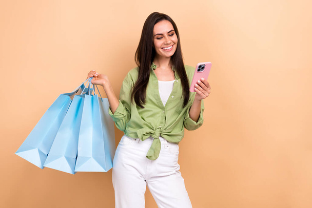 Portrait of gorgeous lovely woman wear green shirt hold new clothes make online order in smartphone isolated on beige color background. - 写真・画像