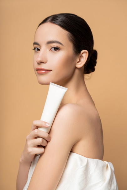 Pretty young asian model in top holding cosmetic lotion and looking at camera isolated on beige - Fotografie, Obrázek