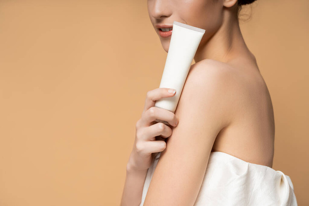 Cropped view of young smiling woman in top holding cosmetic lotion in tube isolated on beige - Fotografie, Obrázek