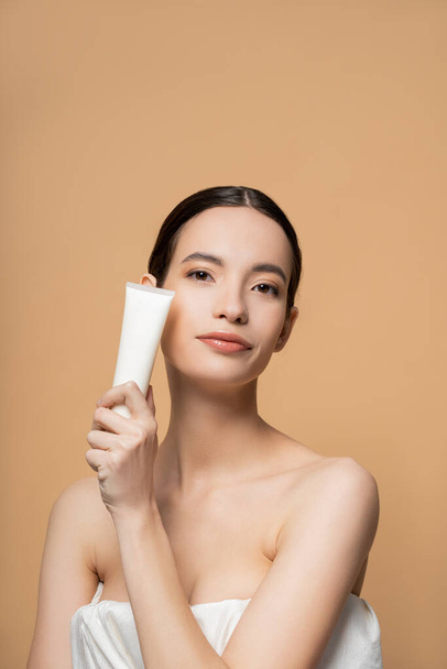 Portrét mladého asijského modelu v horní části držící kosmetický balzám a při pohledu na kameru izolované na béžové - Fotografie, Obrázek
