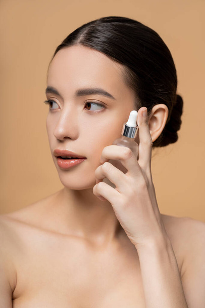 Pretty asian woman with naked shoulders holding cosmetic serum while posing isolated on beige - Photo, Image