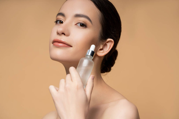Pleased young asian woman with naked shoulders holding cosmetic serum isolated on beige - Фото, зображення