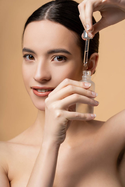 Portrait of confident and smiling asian woman with naked shoulders holding serum isolated on beige - Photo, Image