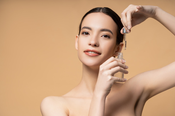 Positive asian woman with naked shoulders posing with cosmetic serum isolated on beige - Foto, afbeelding