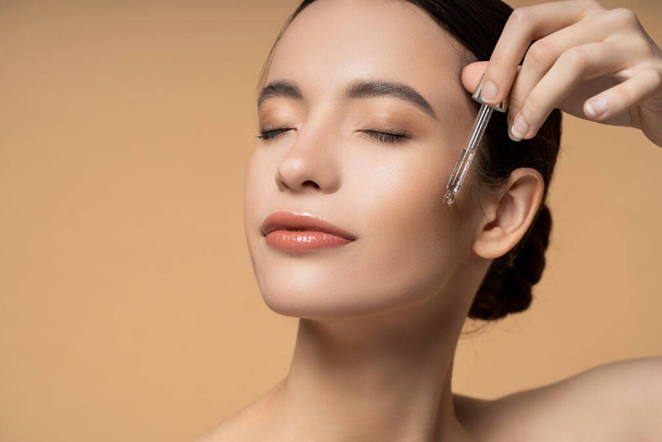 Young asian woman with closed eyes applying cosmetic serum while posing isolated on beige - Φωτογραφία, εικόνα