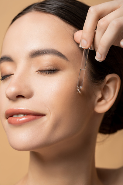 Close up view of cheerful young asian model applying cosmetic serum isolated on beige - Fotó, kép