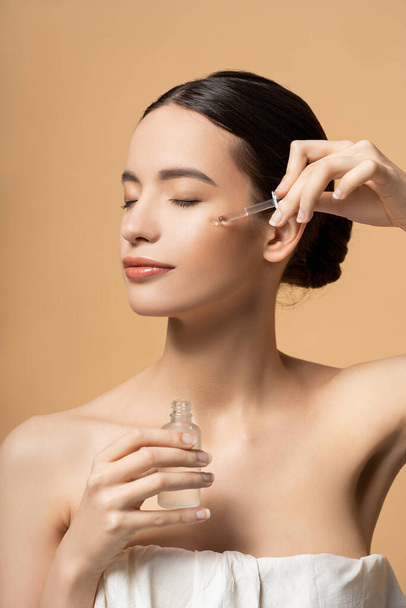 Pretty asian woman with closed eyes in top applying cosmetic serum while posing isolated on beige - Foto, afbeelding