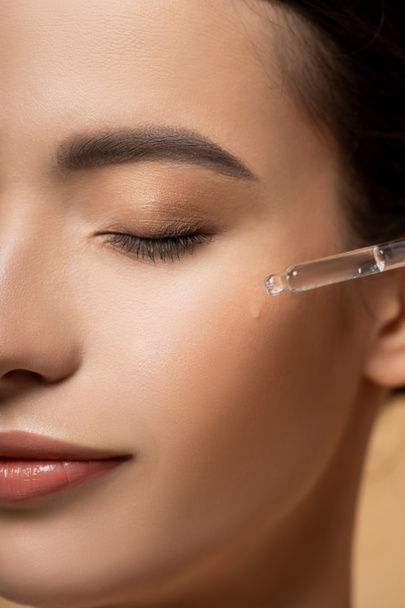 Cropped view of young asian woman applying cosmetic serum on cheek while standing isolated on beige - Foto, afbeelding