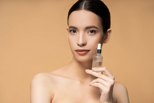 Pretty asian woman with naked shoulders holding cosmetic serum and standing isolated on beige - Valokuva, kuva