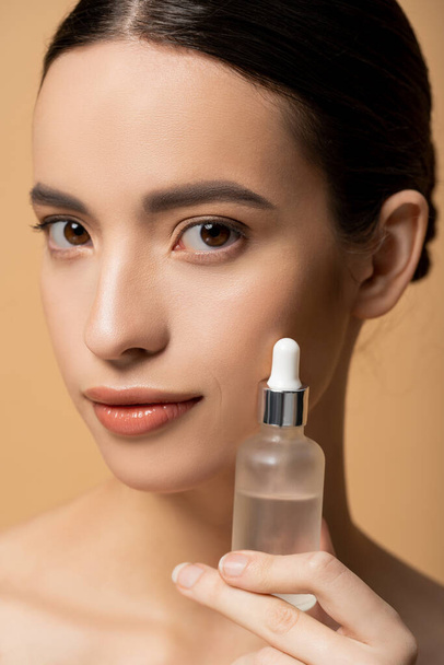 Close up view of young asian woman with perfect skin holding cosmetic serum isolated on beige - Photo, Image