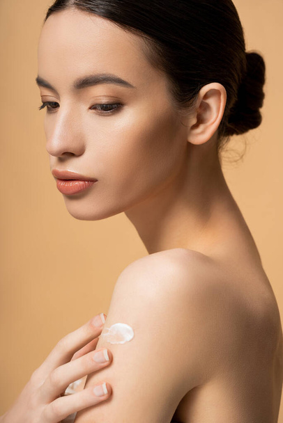 Portrait of young asian brunette woman applying cosmetic cream on shoulder isolated on beige - Фото, изображение
