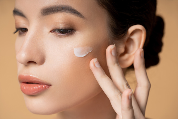 Close up view of young asian woman applying face cream on cheek isolated on beige - Fotografie, Obrázek