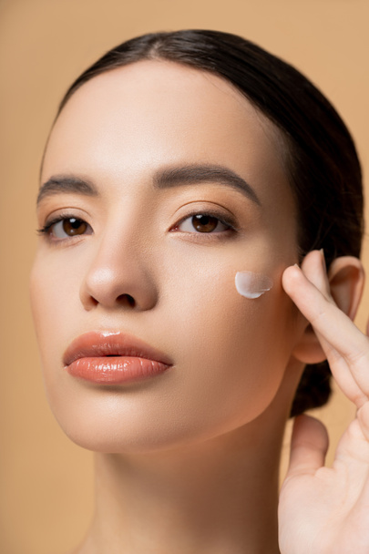 Portrait of young asian woman applying face cream on cheek and looking at camera isolated on beige - Foto, imagen