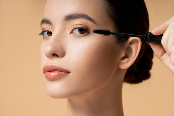 Close up view of young asian woman applying mascara and looking at camera isolated on beige - Foto, Bild