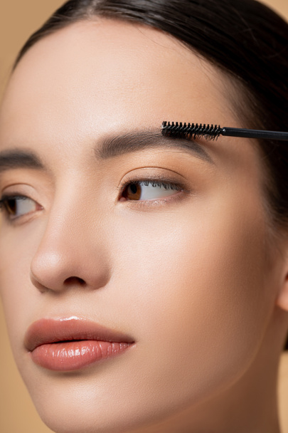Close up view of young asian woman with natural makeup applying eyebrow gel isolated on beige - 写真・画像