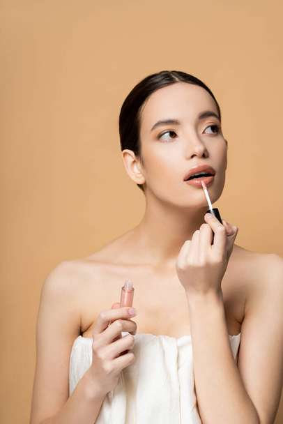 Young brunette asian woman in top applying lip gloss while posing isolated on beige - Foto, afbeelding