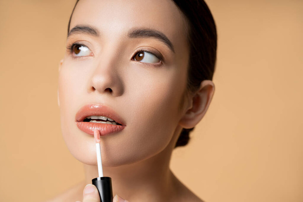 Retrato de joven modelo asiático aplicando brillo de labios y mirando hacia otro lado aislado en beige - Foto, Imagen