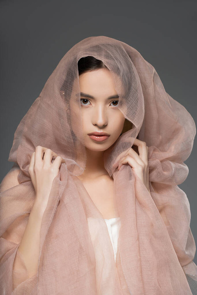 Young asian woman with natural makeup touching beige cloth and looking at camera isolated on grey - Foto, Imagem
