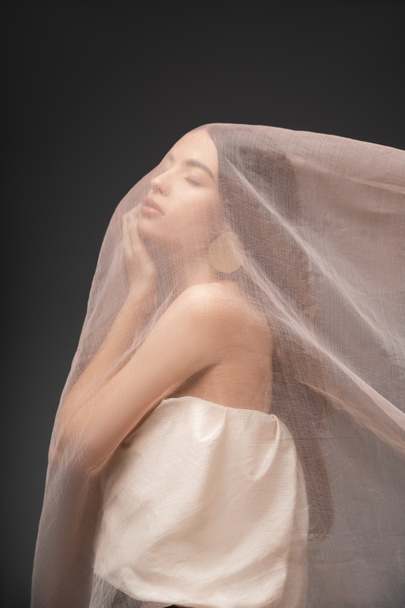 Young asian woman in top touching cheek while posing under beige cloth isolated on grey - Foto, Imagem
