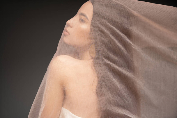 Side view of asian model with naked shoulder looking away while posing under cloth isolated on black - Valokuva, kuva