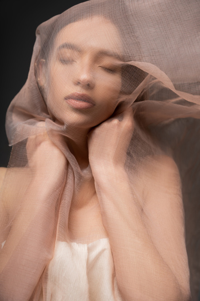 Young asian woman with closed eyes touching beige fabric while posing isolated on black - Zdjęcie, obraz