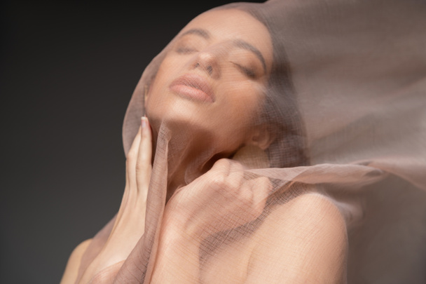 Young asian woman with closed eyes touching neck and posing under beige cloth isolated on black - Fotoğraf, Görsel