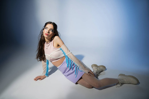 Trendy brunette asian woman in mesh top and skirt looking away while sitting on blue background - Φωτογραφία, εικόνα