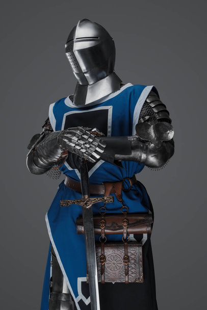 Un soldado medieval vestido con un abrigo azul y una armadura sosteniendo una espada con una pose fija, similar a una estatua, sobre un fondo gris - Foto, Imagen