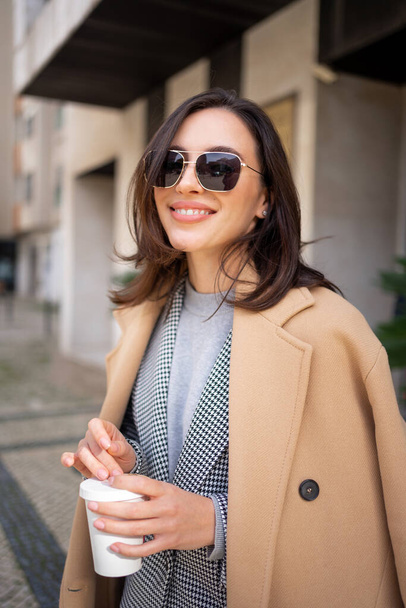 Streetstyle, street fashion concept: woman wearing trendy outfit walking in city. Cream trench coat, sunglasses. Looking camera, vertical photo - Фото, изображение