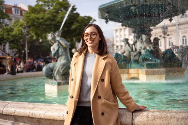 Female tourist enjoying in Lisbon downtown, Portugal. Sunny day. Travel Europe concept. woman dressed stylish coat sitting in front of baroque fountains on Rossio Square, Lisbon. Looking at camera - 写真・画像
