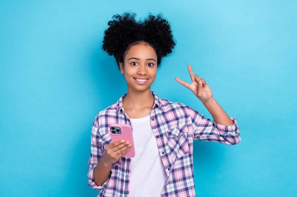 Photo of pleasant girl with buns hairstyle dressed checkered shirt showing v-sign hold smartphone isolated on blue color background. - Photo, image